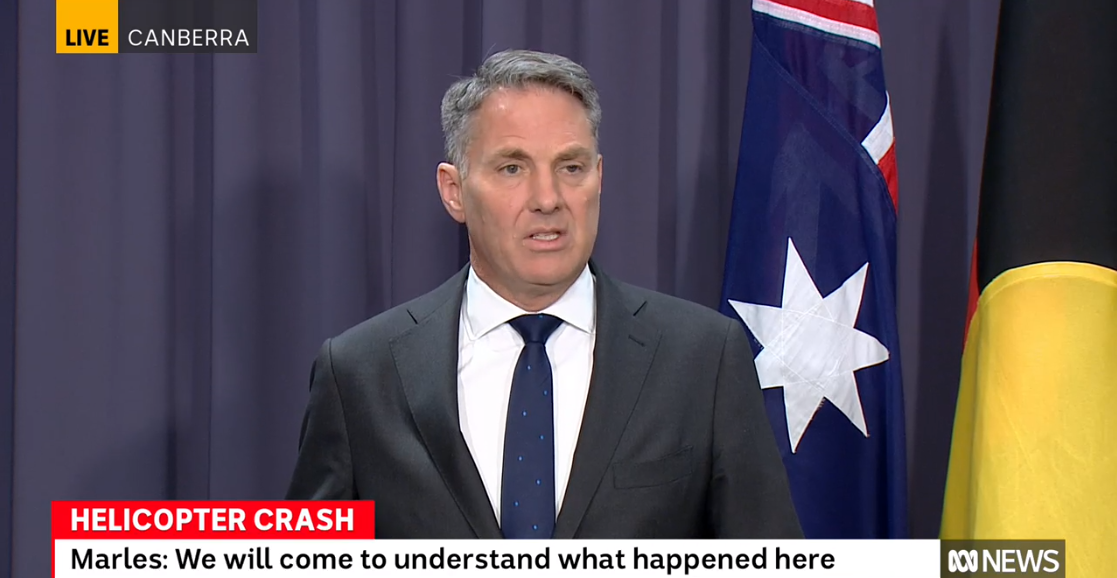 A white man in a suit speaking in front of an Australian flag